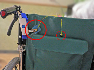 車椅子の背に取り付けられたワイヤレスアラームの送信機に洗濯バサミセンサが接続され、挟んだ絶縁体のカードに結ばれた紐の先にダブルクリップが付いている写真