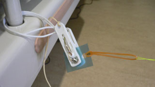 ベッドのフレームに固定された洗濯バサミセンサに絶縁体のカードが挟められており、カードから輪ゴムを介して紐が結ばれている写真